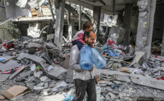 Man carrying child and water bottles across rubble in Gaza