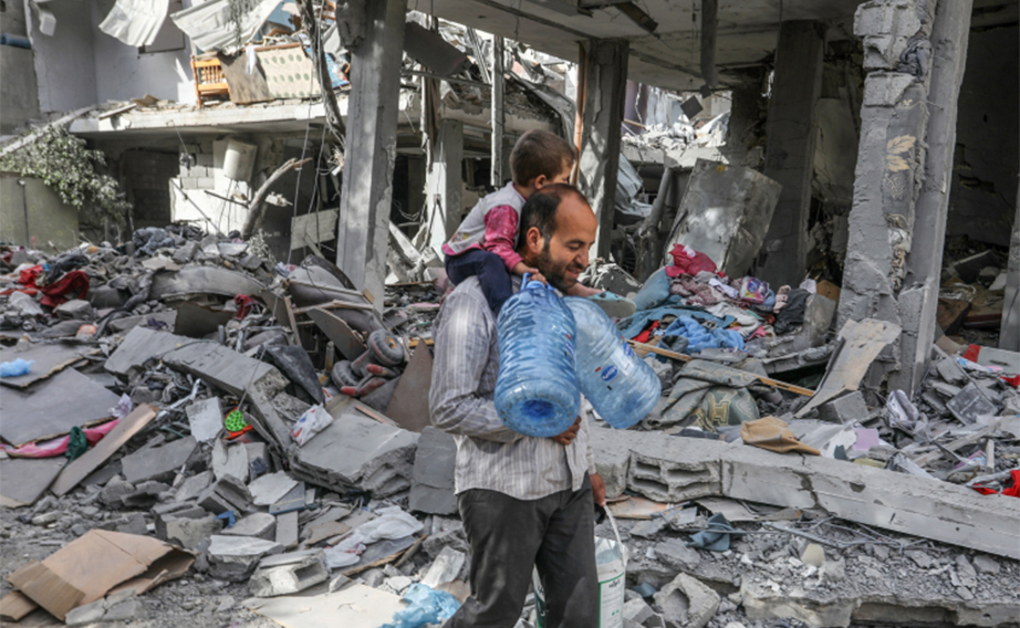 Man carrying child and water bottles across rubble in Gaza